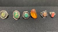 Full view of six vintage men's silver tone rings lined up, showing various green and orange stones and decorative silver-tone settings