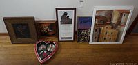Six pieces of framed art displayed on floor including a heart-shaped clock with red and silver tones, floral framed print, poem or quote with woman image, and painting of houses.