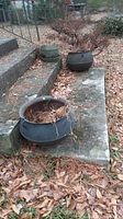 Overview of two cast iron cauldrons on concrete step surrounded by leaves outdoors, showing overall condition and size.