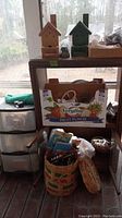 View of 3-drawer plastic storage unit on wheels with labeled boxes and craft supplies on and around a wooden stand, including two birdhouses