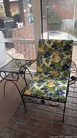 Full view of metal framed chair with floral cushions showing seat and backrest on brick floor porch.