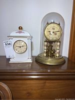 Two clocks on wooden surface, one anniversary clock with glass dome and floral dial, one white decorative mantel clock