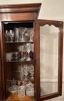 Display cabinet with three shelves showing an assortment of glassware and cut glass containers, including wine glasses, candy dishes, and a biscuit jar.