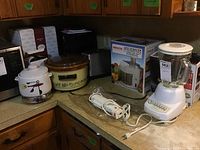 Kitchen countertop holding several kitchen appliances including a white Hamilton Beach blender, Rival crock pot, West Bend bean pot, toaster, boxed Healthmaster juice extractor and George Foreman grill.