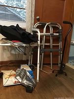 Photo showing two adjustable aluminum walkers, an adjustable quad cane with four feet base and black handle, portable potty seat with folding legs, and various boxes and supplies on the floor.