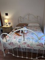 Photo showing full bed setup including white metal bed frame with decorative headboard and footboard, floral bedding and pillows.