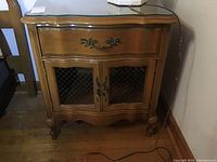 Full front view of wooden nightstand with glass top and closed drawer and doors, sitting on wooden floor.