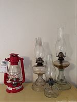 All four lamps shown together on table against wall. One red metal lantern and three clear glass oil lamps of different sizes.