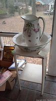 Photo showing the ceramic pitcher and basin on the white wood stand by a window, with visible floral pattern on the pitcher and basin.