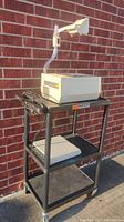 3M overhead projector placed on a black three-tier rolling cart outdoors against a brick wall. Projector is beige with lens arm extended upwards. Power cord coiled beside the projector.