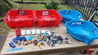 Four Beyblade stadiums (2 red, 2 blue) arranged on a table with Beyblade tops, launchers, and accessories spread in front of them.
