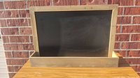 Full view of the rectangular chalkboard with attached wooden tool holder base, placed against a brick wall on a wooden surface.