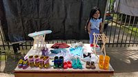 Full view of doll standing on white stand next to wooden ironing board, laundry basket, multiple pairs of doll shoes and assorted clothes arranged on cloth.