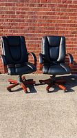 Two black executive office chairs pictured outside against a brick wall, showing front view, padded seat and backrest, wooden armrests, and five-star wooden base with casters.