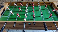 Top view showing red and white player figures on metal rods over a green field with white markings on wooden table.