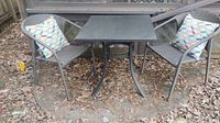 Full view of the patio set with table, two woven chairs, and two accent pillows outdoors on a leaf-covered concrete area.
