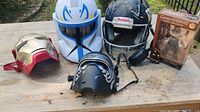 Lot of five items on wooden table showing white storm trooper helmet, black Riddell football helmet, Iron Man mask, black Star Wars mask, and boxed Khal Drogo action figure.