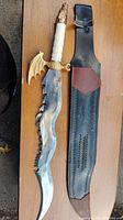 Sword front side with gold dragon handle, white grip, uniquely shaped serrated blade, placed next to black leather sheath with brown accents on a wooden surface.