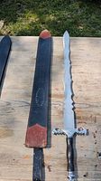 Silver sword with wavy blade and ornate crossguard alongside black leather sheath on a wooden surface in daylight.