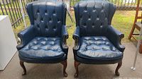 Front view of both navy blue leather wingback chairs side by side showing the deep button tufting, rolled arms with nailhead trim, and wooden cabriole front legs.