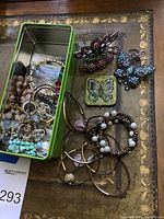 Green tin box opened on table showing assorted bracelets, rings, earrings, brooches