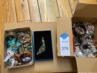 Overview of two cardboard boxes containing assorted jewelry on wooden floor