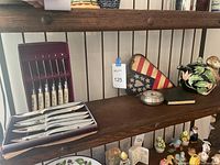 Full shelf showing Royal Brand cutlery set in box, ceramic monkey teapot, small metal container, patriotic heart decoration, and other items.