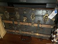 Front view of vintage wooden storage chest showing metal latches and leather straps, with measurement tag attached.