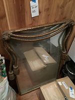 Mirror showing metal frame with rope and tassel design, placed leaning against a wooden wall with cardboard boxes around it.