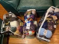 Three large clear bags packed with assorted yarn in various sizes, colors and textures on wooden floor, with dark green sofa in background.