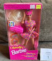 Front view of unopened pink box displaying Barbie doll in floral dress holding pink Caboodles makeup case and wearing a pink cap.
