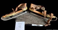 Pair of early 1900s antique skates showing wooden construction, metal blade, and leather straps horizontally on a dark surface with Sharpie and MaxSold paper for scale.