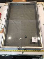 Top-down view showing the full glass lid with metal frame and cloth insert inside. Two small metal objects and a chain-link are visible inside the case.