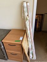 Wooden two-drawer file cabinet with metal handles, wooden foldable TV tray leaning against it, and two bolts of floral fabric standing upright next to the cabinet.