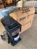 BUNN coffee maker with glass carafe next to original BUNN box, placed on carpeted floor