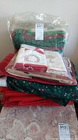 Stack of folded Christmas-themed tablecloths and linens, showing various colors and patterns including red, green with holly print, and white with embroidered wreath.