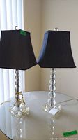 Pair of glass ball base electric table lamps with black shades on a round glass table near vertical blinds, showing overall design