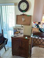 Showcases the slow cooker in original box on top of the wooden storage cabinet, with round black-framed wall clock and rectangular cork bulletin board mounted on wall above.
