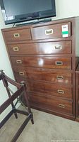 Full front view of tall wood dresser showing two smaller top drawers and five long lower drawers with recessed gold-tone metal handles. Dust and light scratches visible on surface.
