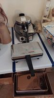Photo showing a stainless steel Waring Pro pizzelle baker and a rectangular Villa Ware waffle maker with black handle, both placed on a table.
