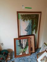 Four framed artworks leaning against a wall: two paintings on canvas in wooden frames, two framed prints partially visible stacked on the floor. Paintings depict natural landscapes with snow and mountains.