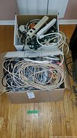 Two boxes on floor filled with assorted cables, extension cords, and power strips visibly tangled together.