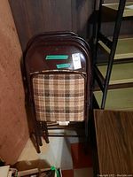 Four folding chairs stacked against wall, showing metal frames and plaid upholstered padded seats and backs.