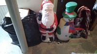 Lighted plastic Santa Claus and snowman figures, two lighted candy canes leaning against dark wood panel, plastic bags in background