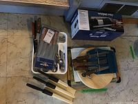 Photo showing knives with wooden handles, assorted forks and spoons in white tray, commercial cutter with multiple wooden handled blades on wooden cutting board, sharpener box on floor.