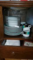 Side view of stacked dinnerware showing plate edges and cups with Midnight Emerald striping inside a wooden cabinet.