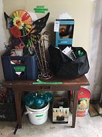 Overview photo of garden supplies including metal stakes, plastic crate with items, black bags of bulbs, white bucket, and boxed light fixture on a wooden table.