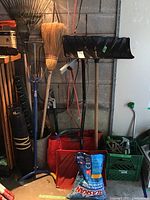 Photo of garden tools including snow shovels, broom, rakes, and leaf bags arranged against a concrete wall.