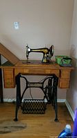 Front-facing photo of the sewing machine mounted in the cabinet with the vintage iron and green basket on top.