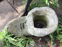 Concrete shoe planter shown outdoors surrounded by grass and plants, displaying worn and mossy surface with a hollow center for planting.
